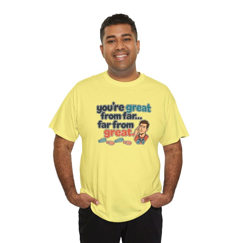 May include: A light yellow short-sleeve t-shirt featuring the text "you're great from far... far from great." in blue and red, accompanied by an illustration of a person looking through binoculars and four small colorful footprints.
