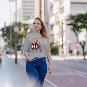May include: A light gray short-sleeve t-shirt featuring a graphic of a woman with long wavy hair, the text "HI Ask me anything..." and small icons representing chat, voice, and image functions printed on the front center of the garment.
