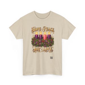 May include: A beige t-shirt featuring a graphic of four lit candles nestled in a green wreath with purple ribbons. The text reads HOPE, PEACE, JOY, and LOVE in gold lettering. A small square QR code is printed near the bottom right of the graphic.