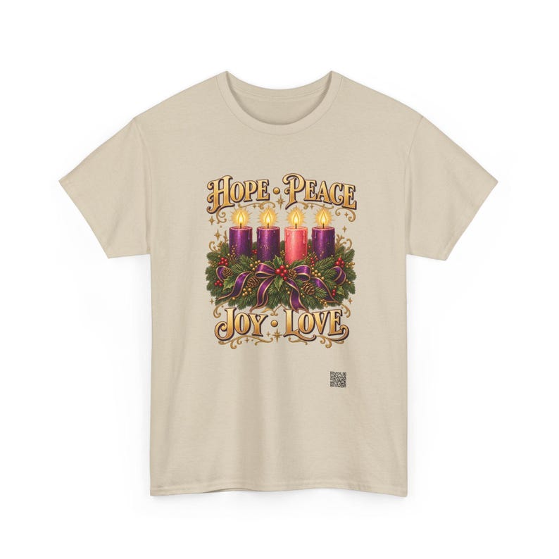 May include: A beige t-shirt featuring a graphic of four lit candles nestled in a green wreath with purple ribbons. The text reads HOPE, PEACE, JOY, and LOVE in gold lettering. A small square QR code is printed near the bottom right of the graphic.