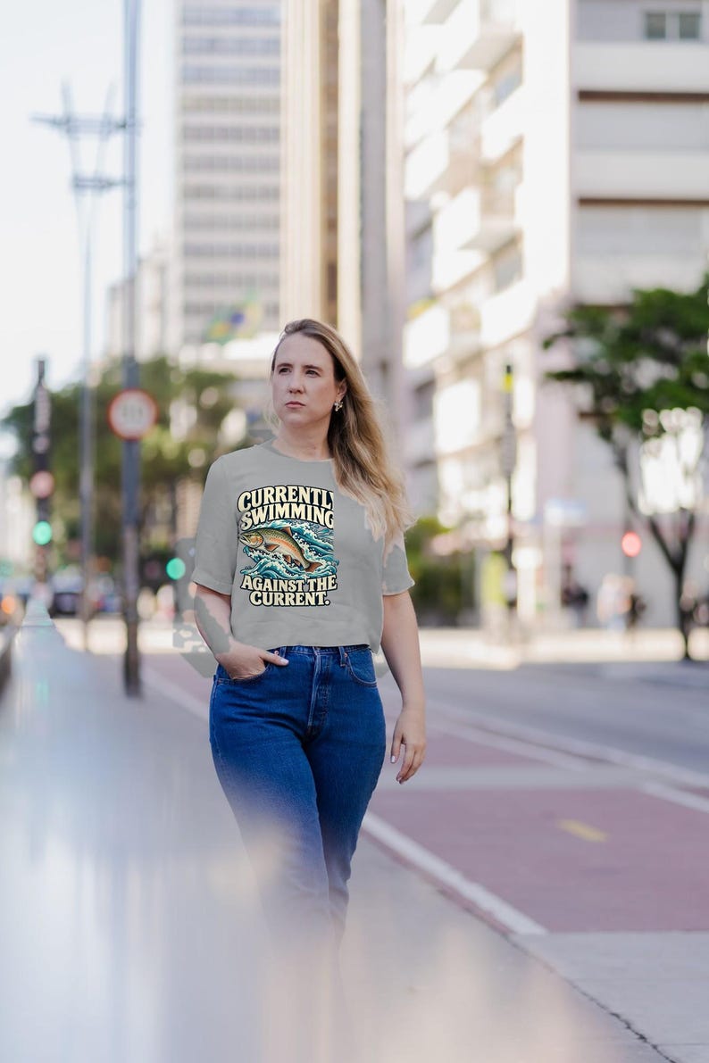 May include: A light gray short-sleeved t-shirt featuring a graphic of a fish jumping through blue waves. The text on the shirt reads "CURRENTLY SWIMMING AGAINST THE CURRENT." The shirt has a crew neckline and a straight hemline.