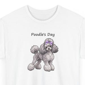 May include: A white t-shirt featuring a graphic illustration of a grey poodle with a purple bow on its head. Above the dog, the text Poodle's Day is printed in a simple black font. The poodle has curly fur and is standing in profile.