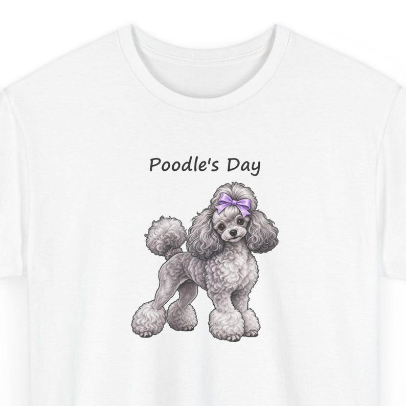 May include: A white t-shirt featuring a graphic illustration of a grey poodle with a purple bow on its head. Above the dog, the text Poodle's Day is printed in a simple black font. The poodle has curly fur and is standing in profile.