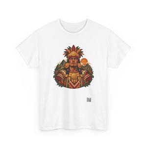 May include: A white t-shirt featuring a central graphic of a person wearing a feathered headdress and ornate gold jewelry. The design includes mountain silhouettes, a sun, and geometric patterns in red, green, and gold tones. A small QR code is visible below.