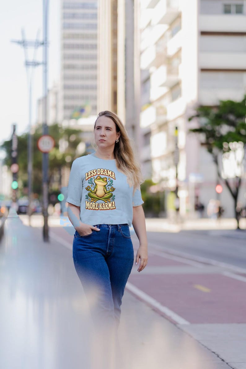 May include: A light blue short-sleeve t-shirt featuring a graphic of a green frog sitting in a lotus position. The text above the frog reads LESS DRAMA and the text below reads MORE KARMA, surrounded by small blue star and swirl accents.