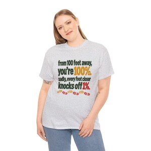 May include: A light gray short-sleeve t-shirt featuring the black text: from 100 feet away, you're 100%. sadly, every foot closer knocks off 1%. The text includes the number 100% in gold and 1% in red, with small colorful footprints below.