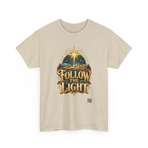 May include: A beige short-sleeve t-shirt featuring a graphic print of a glowing star above a landscape with the text FOLLOW THE LIGHT in gold lettering. The design includes stylized hills, a small town, and twinkling stars in a dark blue sky.
