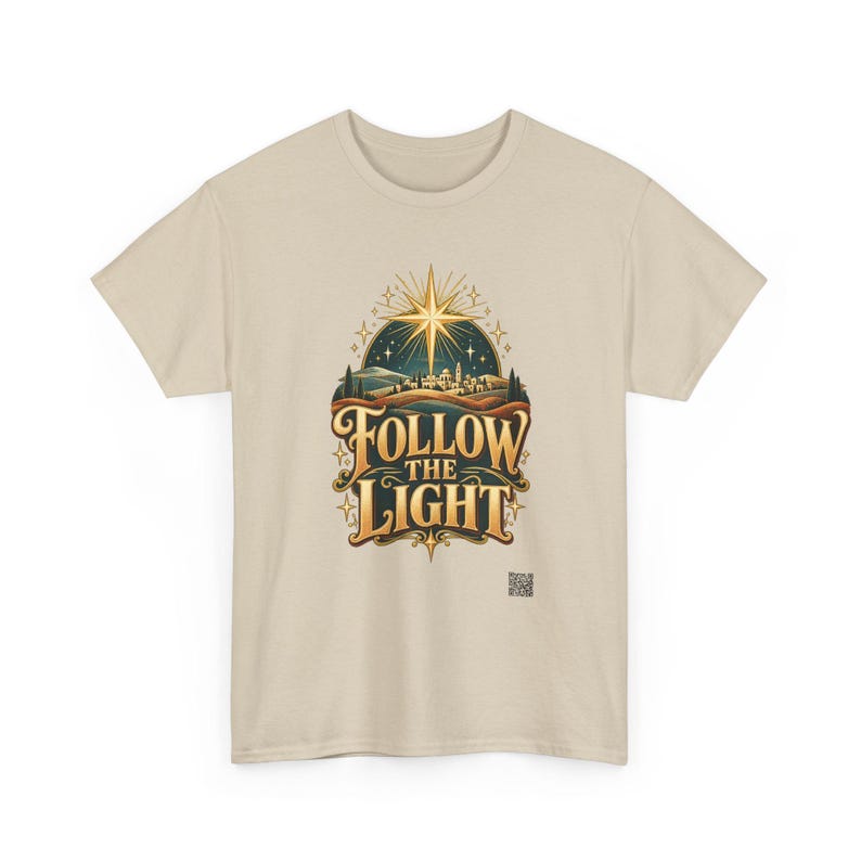 May include: A beige short-sleeve t-shirt featuring a graphic print of a glowing star above a landscape with the text FOLLOW THE LIGHT in gold lettering. The design includes stylized hills, a small town, and twinkling stars in a dark blue sky.