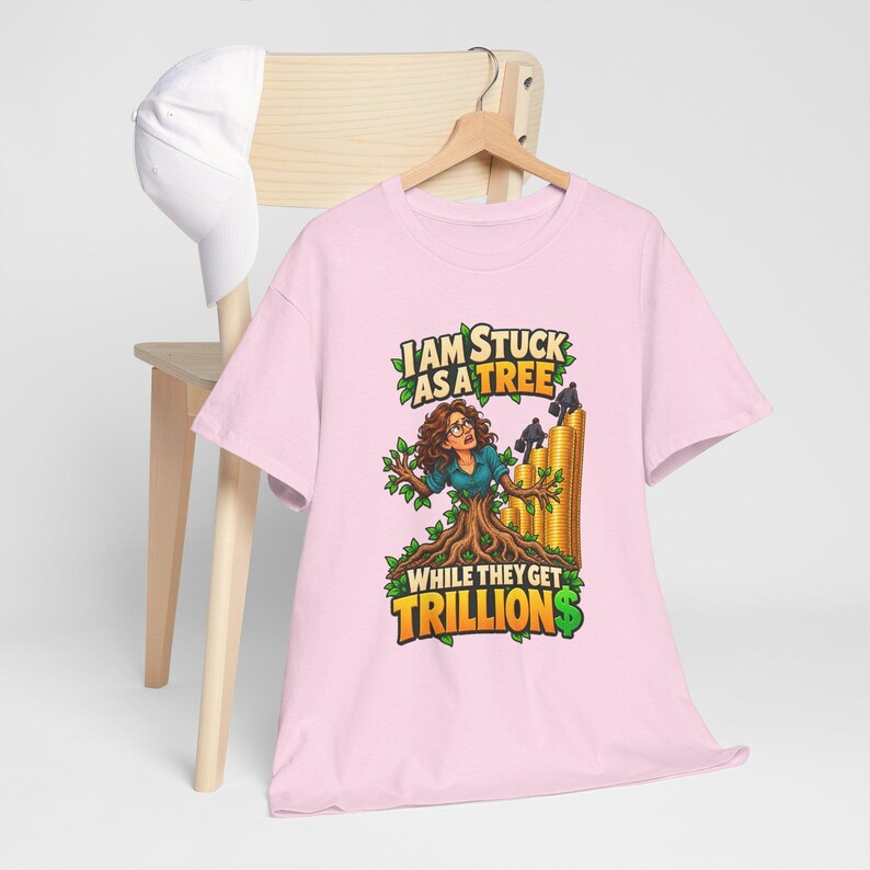 May include: A light pink t-shirt featuring a graphic illustration of a person merged with a tree trunk, surrounded by green leaves and gold coin stacks. The text reads I AM STUCK AS A TREE WHILE THEY GET TRILLIONS in bold yellow and green letters.
