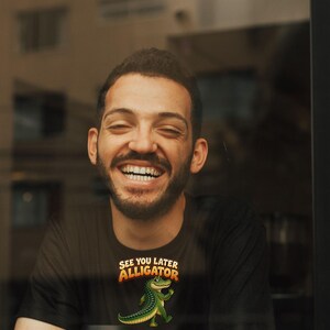 May include: A black short-sleeved t-shirt featuring a graphic of a green alligator waving its hand. Above the alligator, the text reads "SEE YOU LATER ALLIGATOR" in a bold, orange, arched font. The shirt is made of a solid black fabric.