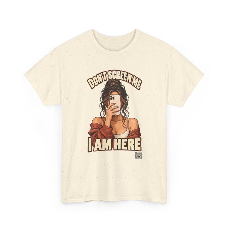 K&ouml;nnte beinhalten: Ein cremefarbenes Kurzarm-T-Shirt mit einer Grafik einer Person, die ein Smartphone vor das Gesicht h&auml;lt. Der Text "DON'T SCREEN ME I AM HERE" ist in Braun &uuml;ber und unter der Illustration gedruckt, die einen kleinen QR-Code enth&auml;lt.