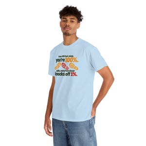 May include: A light blue short-sleeve t-shirt featuring the black text "from 100 feet away, you're 100%." above three colorful shoe print icons, followed by the text "sadly, every foot closer knocks off 1%." in black and red.