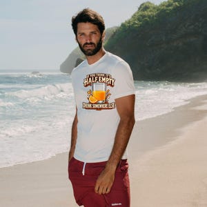 May include: A white short-sleeve t-shirt featuring a graphic of a glass of orange juice with an orange slice and the text "IF YOU THINK IT'S HALF EMPTY DRINK SOMEWHERE ELSE" paired with maroon shorts featuring a white drawstring.