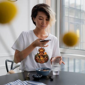 May include: A white short-sleeved t-shirt featuring a colorful graphic print of a figure wearing an elaborate feathered headdress and traditional attire. The design is centered on the chest area of the garment, which is made of a plain white fabric.