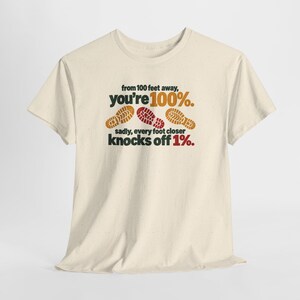 May include: A cream-colored short-sleeve t-shirt featuring the text 'from 100 feet away, you're 100%. sadly, every foot closer knocks off 1%.' with three brown and red shoe print graphics centered on the front of the garment.