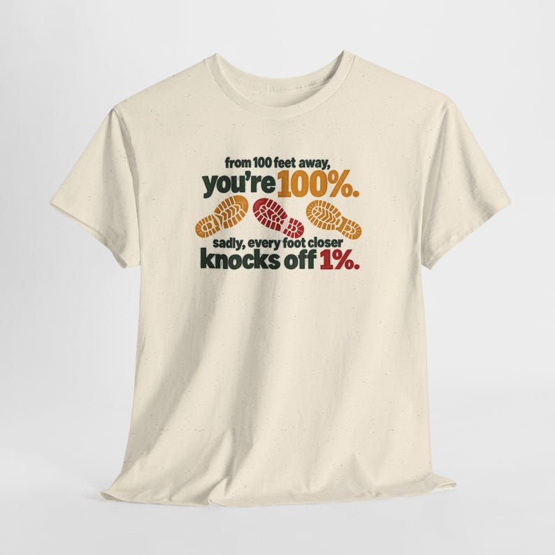 May include: A cream-colored short-sleeve t-shirt featuring the text 'from 100 feet away, you're 100%. sadly, every foot closer knocks off 1%.' with three brown and red shoe print graphics centered on the front of the garment.