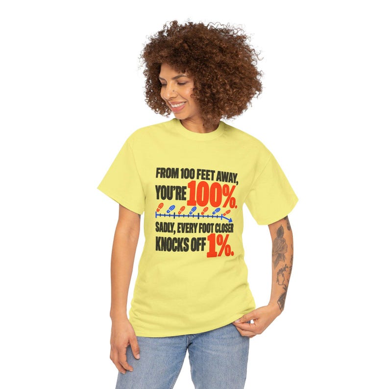 May include: A pale yellow short-sleeve t-shirt featuring black and red text that reads: FROM 100 FEET AWAY, YOU'RE 100%. SADLY, EVERY FOOT CLOSER KNOCKS OFF 1%. The design includes a blue line with small red and blue shoe icons.