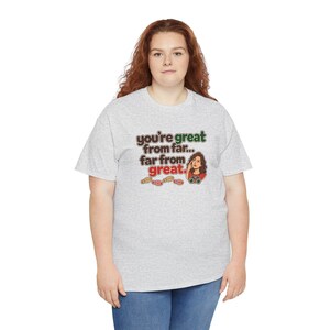 May include: A light gray short-sleeve t-shirt featuring the text "you're great from far... far from great." in brown and green, accompanied by an illustration of a person looking through binoculars and several small footprints.