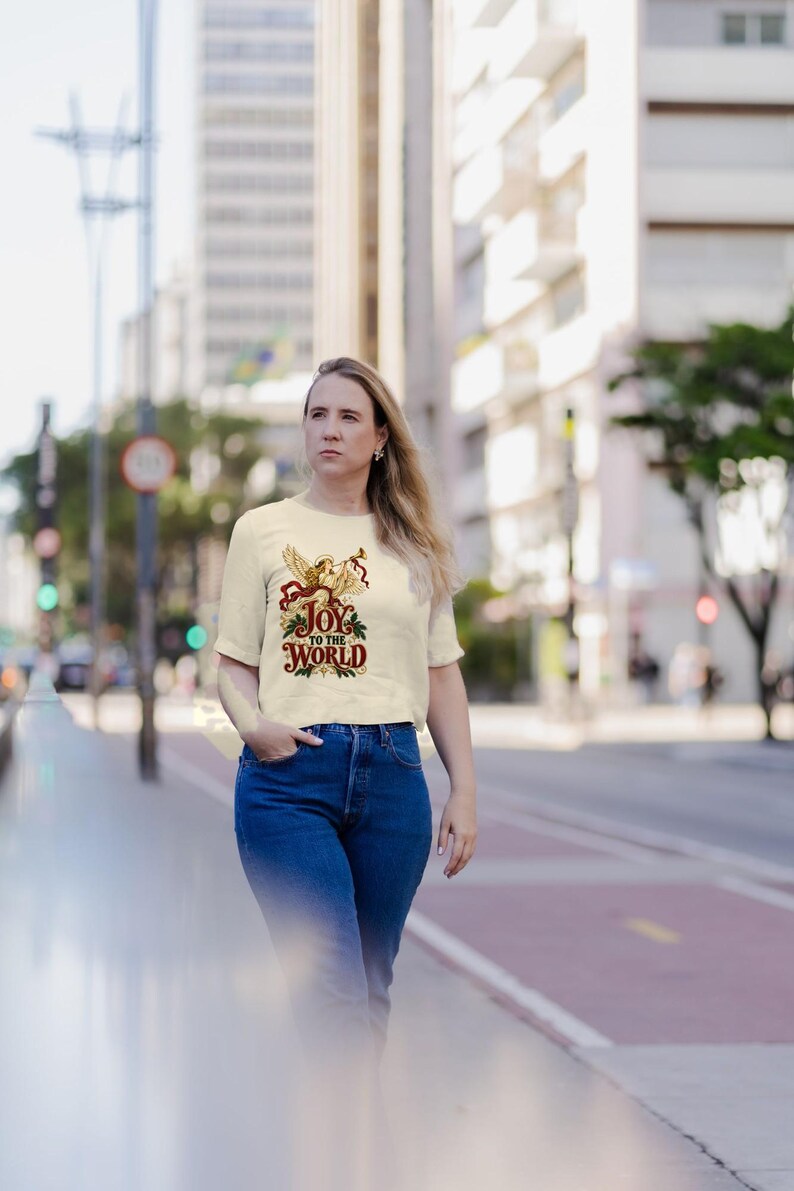 May include: A cream-colored short-sleeve t-shirt featuring a graphic print of a golden angel playing a trumpet, surrounded by red ribbons and green holly leaves, with the text "JOY TO THE WORLD" printed in a decorative red and green font.