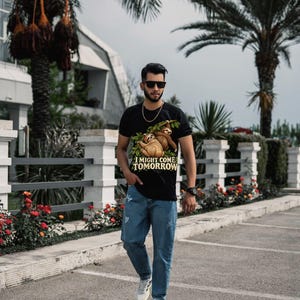 Pode incluir: Uma t-shirt preta com um gr&aacute;fico de duas pregui&ccedil;as penduradas num ramo com o texto I MIGHT COME TOMORROW, combinada com cal&ccedil;as de ganga azuis, uma corrente dourada, &oacute;culos de sol pretos, um rel&oacute;gio preto e sapatilhas brancas.