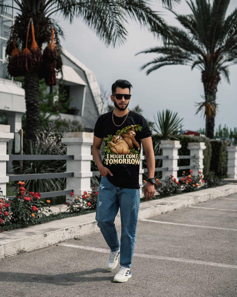 Pode incluir: Uma t-shirt preta com um gr&aacute;fico de duas pregui&ccedil;as penduradas num ramo com o texto I MIGHT COME TOMORROW, combinada com cal&ccedil;as de ganga azuis, uma corrente dourada, &oacute;culos de sol pretos, um rel&oacute;gio preto e sapatilhas brancas.