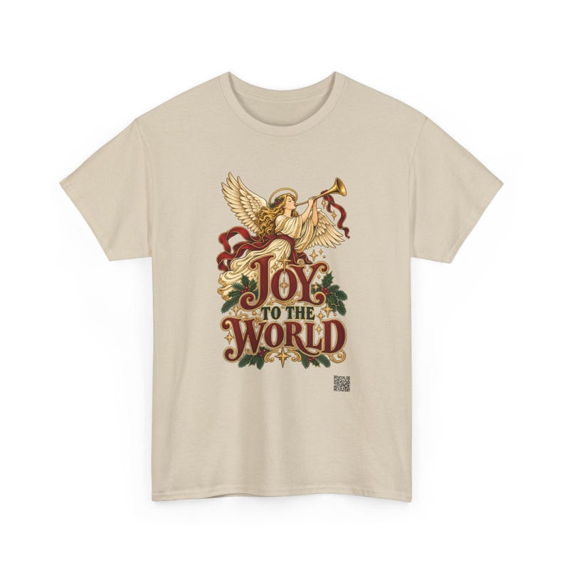 May include: A beige short-sleeved t-shirt featuring a graphic of an angel with wings playing a golden trumpet. Below the angel, the text reads "JOY TO THE WORLD" in red and gold lettering, surrounded by green holly leaves and small stars.