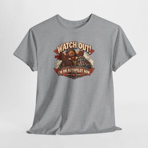 May include: A heather gray short-sleeve t-shirt featuring a graphic of a pilot and a small robot with the text WATCH OUT! I'M ON AUTOPILOT NOW. The design includes a propeller emblem and clouds in a vintage-inspired illustration style.
