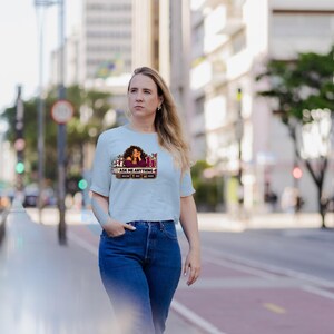 May include: A light blue short-sleeved t-shirt featuring a graphic print of a person with curly hair and the text "ai h" and "ASK ME ANYTHING" with small icons below the text. The shirt is paired with dark blue denim jeans.