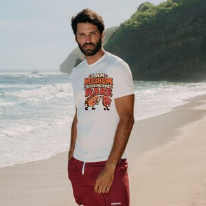 May include: A white short-sleeved t-shirt featuring a graphic print with the text "I AM MEDIUM LOOKING FOR RARE" in orange and red letters, accompanied by cartoon illustrations of a steak piece with a magnifying glass and a heart.