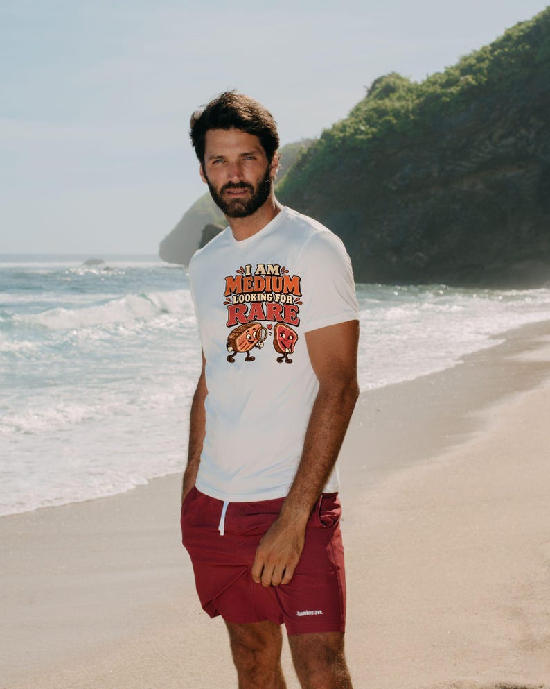 May include: A white short-sleeved t-shirt featuring a graphic print with the text "I AM MEDIUM LOOKING FOR RARE" in orange and red letters, accompanied by cartoon illustrations of a steak piece with a magnifying glass and a heart.