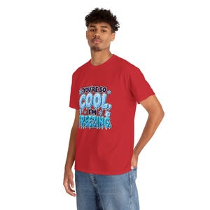May include: A red short-sleeved t-shirt featuring a graphic design with the text "YOU'RE SO COOL, I'M FREEZING." The lettering is styled in a light blue, icy, frozen font, accented with small white snowflakes and ice cube illustrations.