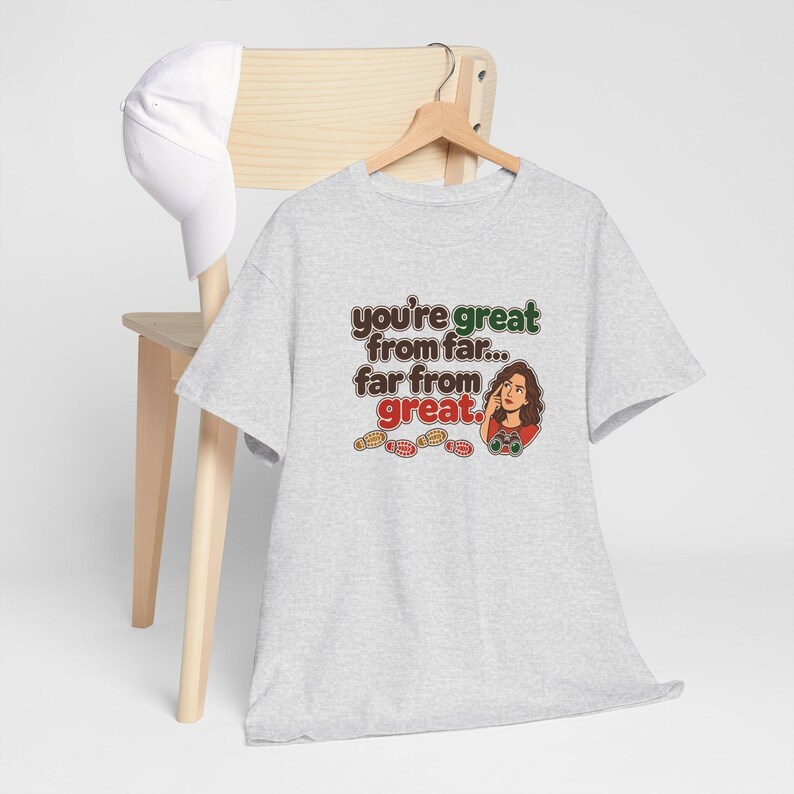May include: A light gray short-sleeve t-shirt featuring a graphic with the text "you're great from far... far from great." The design includes an illustration of a person with binoculars and several small footprints in brown, green, and red.
