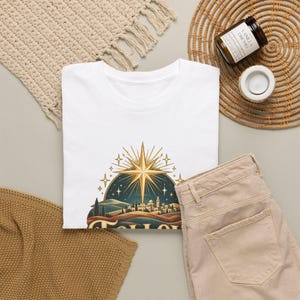 May include: A white short-sleeved t-shirt featuring a graphic print of a golden star shining over a stylized landscape with a small town, rolling hills, and a dark blue night sky filled with smaller stars and light rays.
