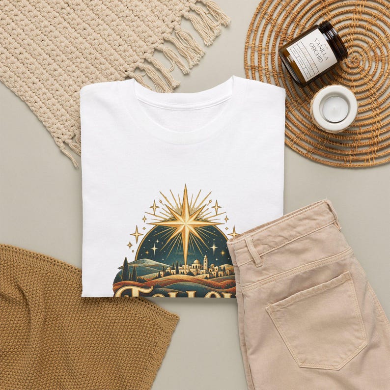 May include: A white short-sleeved t-shirt featuring a graphic print of a golden star shining over a stylized landscape with a small town, rolling hills, and a dark blue night sky filled with smaller stars and light rays.