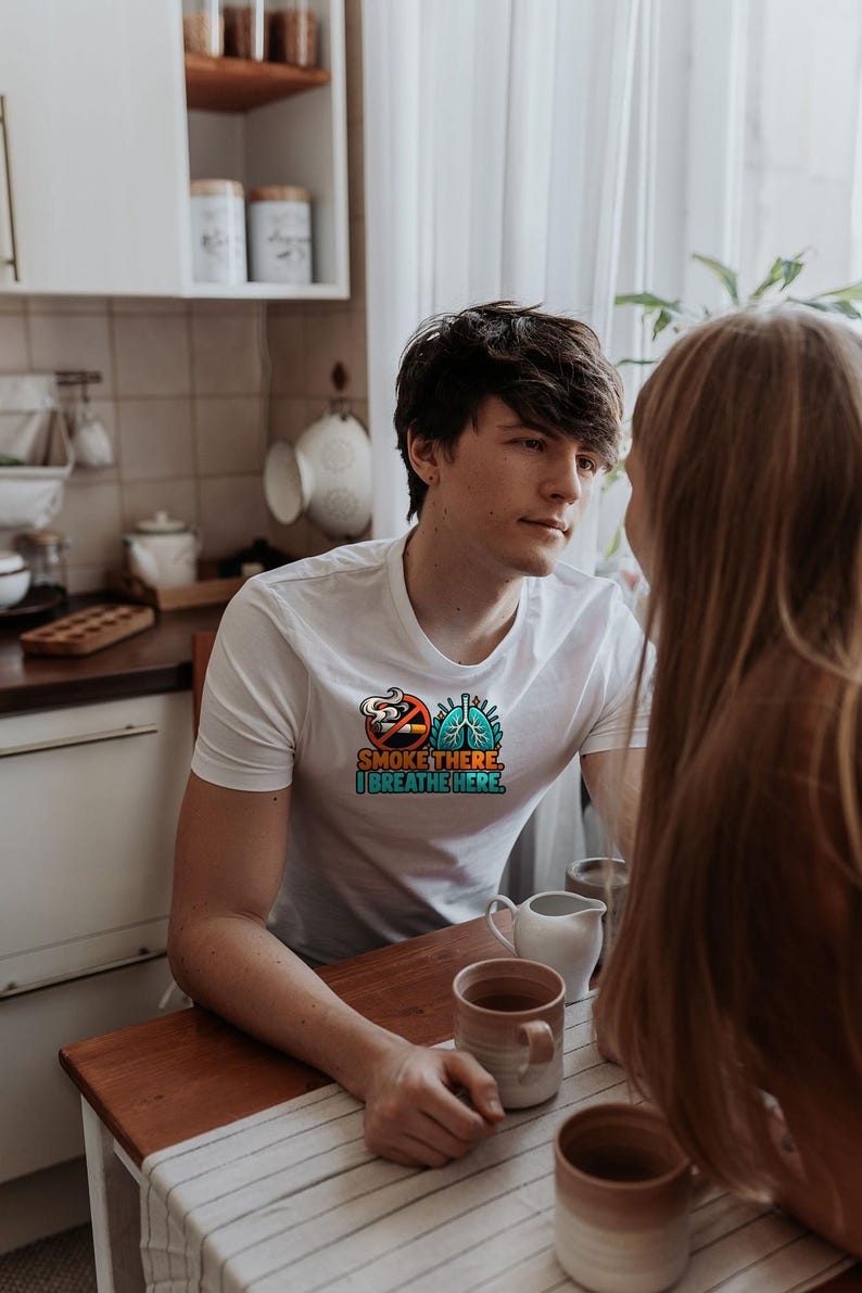 May include: A white short-sleeved t-shirt featuring a graphic print on the chest. The design includes a no-smoking symbol, a pair of blue lungs, and the text "SMOKE THERE. I BREATHE HERE." in orange and teal block lettering.
