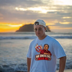 May include: A white t-shirt featuring a graphic print of a man with crossed arms, a no-smoking symbol, and the text "NO SMOKING NEAR ME" in red and white lettering. The design includes a cigarette with a red prohibition circle and smoke.