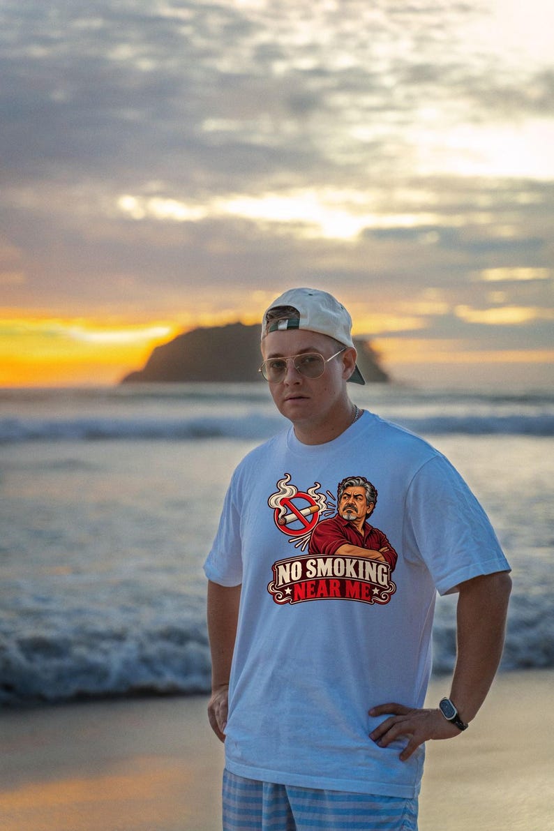 May include: A white t-shirt featuring a graphic print of a man with crossed arms, a no-smoking symbol, and the text "NO SMOKING NEAR ME" in red and white lettering. The design includes a cigarette with a red prohibition circle and smoke.