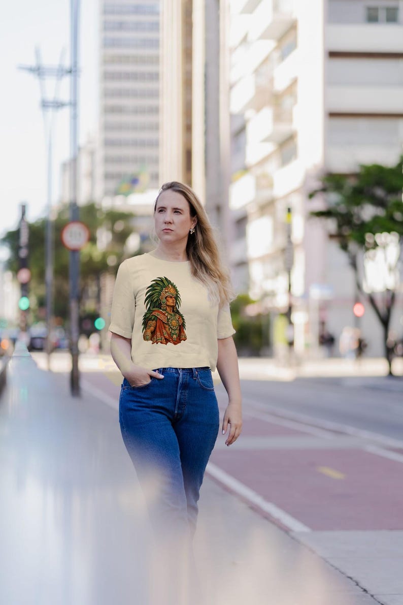 May include: A cream-colored short-sleeve t-shirt featuring a graphic print of an Aztec warrior wearing a large green feathered headdress and traditional orange and gold attire, paired with dark blue denim jeans.