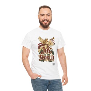 May include: A white short-sleeved t-shirt featuring a graphic of an angel playing a trumpet with red ribbons and green holly leaves. Below the angel, the text reads JOY TO THE WORLD in large, stylized brown letters with gold accents.