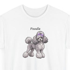 May include: A white t-shirt featuring a graphic illustration of a gray poodle with a purple bow on its head. The word Poodle is printed in black text above the dog. The poodle has curly fur and is standing in a profile position.