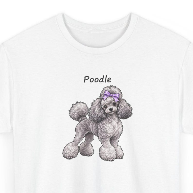 May include: A white t-shirt featuring a graphic illustration of a gray poodle with a purple bow on its head. The word Poodle is printed in black text above the dog. The poodle has curly fur and is standing in a profile position.