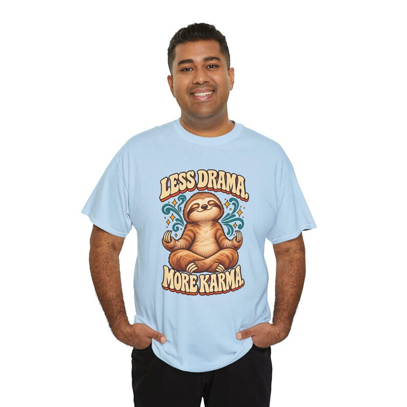 May include: A light blue short-sleeve t-shirt featuring a graphic of a brown sloth sitting in a lotus position. The text above the sloth reads LESS DRAMA and the text below reads MORE KARMA. The design includes teal swirls and small yellow stars.