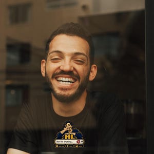 May include: A black short-sleeved t-shirt featuring a graphic of a person with glasses resting their chin on their hand above the text HI and Ask me anything, with icons for new chat, voice, and image, plus a white coffee cup with a black logo.