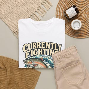 May include: A white t-shirt featuring a graphic of a trout jumping out of blue waves with the text "CURRENTLY FIGHTING" in large, bold, cream-colored letters with a dark outline, positioned above the fish illustration.