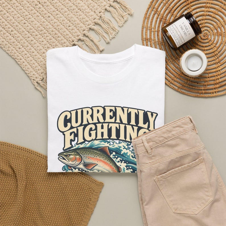 May include: A white t-shirt featuring a graphic of a trout jumping out of blue waves with the text "CURRENTLY FIGHTING" in large, bold, cream-colored letters with a dark outline, positioned above the fish illustration.