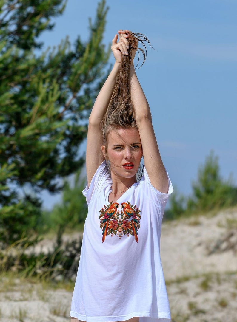 May include: A white short-sleeved t-shirt featuring a vibrant, symmetrical graphic print on the chest. The design displays two colorful parrots facing each other, surrounded by detailed tropical flowers and foliage in shades of red, orange, and green.