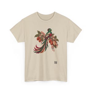 May include: A beige t-shirt featuring a central graphic of a quetzal bird perched on a branch with cacao pods and leaves. The bird has green, red, and white plumage with a long tail. Geometric patterns and a small QR code are visible.