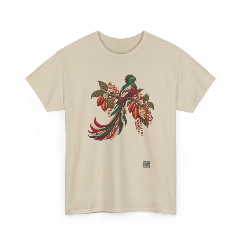 May include: A beige t-shirt featuring a central graphic of a quetzal bird perched on a branch with cacao pods and leaves. The bird has green, red, and white plumage with a long tail. Geometric patterns and a small QR code are visible.