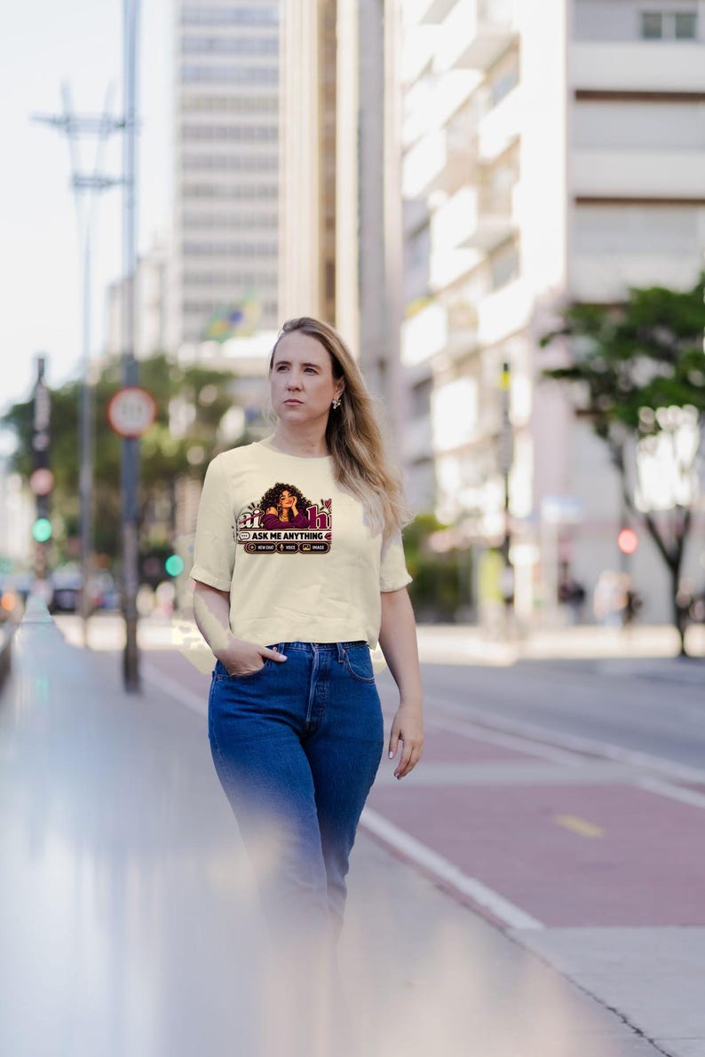 May include: A cream-colored short-sleeve t-shirt featuring a graphic print of a person with dark curly hair, the text "Ask Me Anything" in a stylized font, and smaller icons arranged in a horizontal row below the main text.