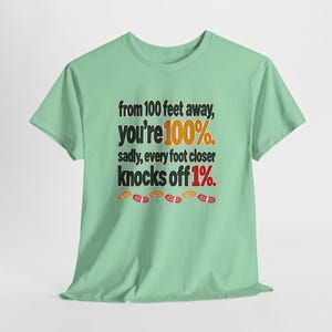 May include: A light green short-sleeve t-shirt featuring the black text: from 100 feet away, you're 100%. sadly, every foot closer knocks off 1%. The text includes orange and red accents, with small orange and red shoe print icons below.